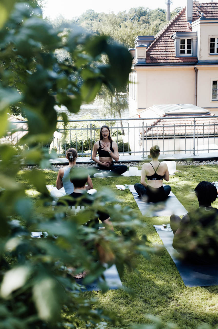 Participants seated on mats on a lakeside lawn during a guided community practice in Berlin.